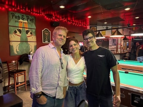 Three people posing together inside a bar with red lighting, pool tables, and a Pug and Pug Brewing sign visible