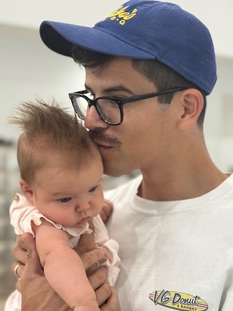 Man wearing blue cap and glasses holding a baby in pink outfit indoors