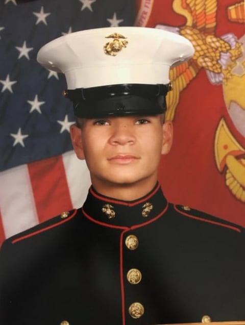 U.S. Marine in full dress uniform with white cover and black dress uniform, American flag in background