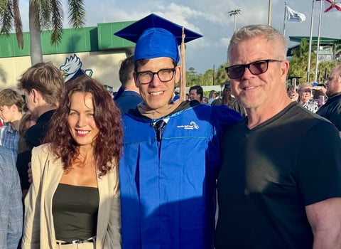 Graduate in blue cap and gown standing between two adults at an outdoor graduation ceremony with palm trees and flags in background