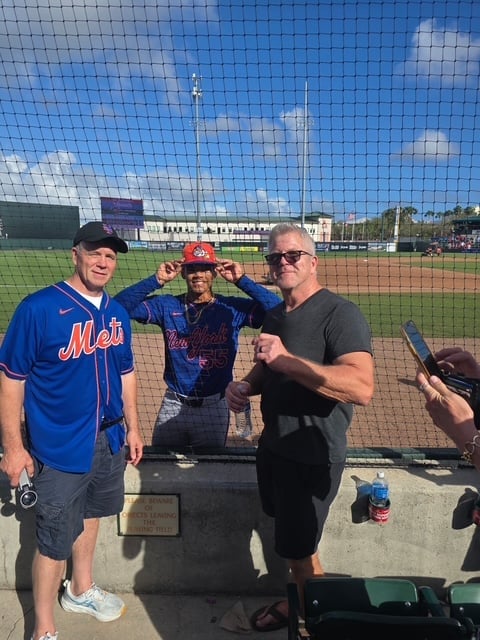 Three men posing on a baseball field with backstop netting, one wearing a Mets jersey and another in Cardinals attire, with city skyline visible in background